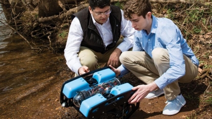 Two men crouched around a robot along a river bank