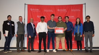 Group of students standingwith professors and judges in front of a step and repeat banner with the center three holding their first place certificate.