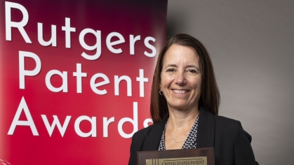 Woman wearing a black and white dress and black blazer holds award plaque in front of a red banner that says Rutgers Patent Awards.of a 