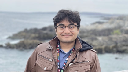 Male engineering student headshot posing outdoors with water and a rocky coastline behind him. He has dark hair, glasses and is wearing a brown jacket over a blue shirt.