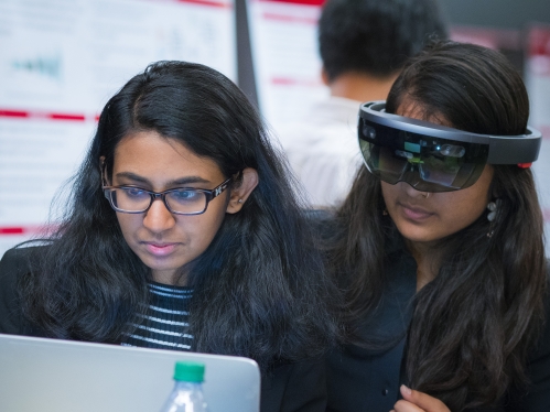 One female with long black hair wearing a virtual reality headset and another female with long hair with eyeglasses looking at a computer screen.