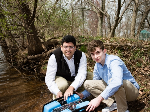 Male professor and male student squatting next to an underwater acoustic communications vehicle on the river bank.