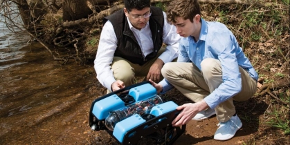 Two men crouched around a robot along a river bank