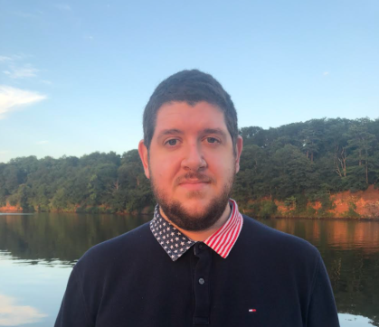 Head shot of male wearing a dark blue sweater with red, white and blue stars and striped collar standing with a lake in the background.