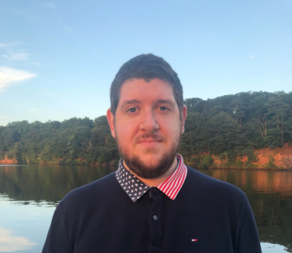 Head shot of male wearing a dark blue sweater with red, white and blue stars and striped collar standing with a lake in the background. 