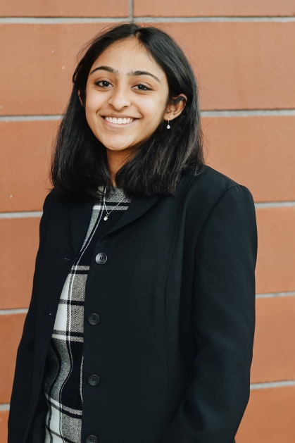 Young woman poses in front of a brick wall. She has black hair and is wearing a black jacket.