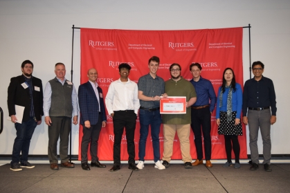 Group of students standingwith professors and judges in front of a step and repeat banner with the center three holding their first place certificate.
