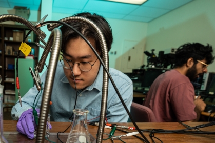Male student wearing protective gloves studies contents of beaker connected to a circuit