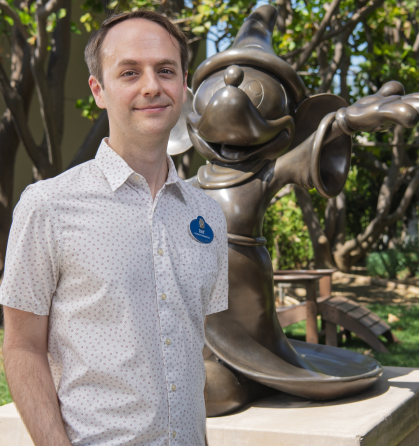 Man with receding hairline wearing a white shirt and standing next to a sculpture of Mickey Mouse