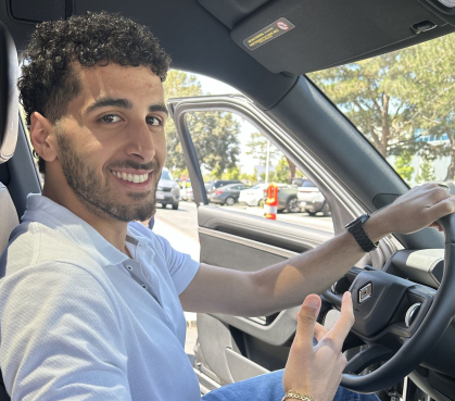 Man with dark hair, a beard and mustache wearing white short-sleeved shirt sitting in a car holding the steering wheel