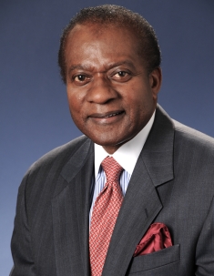 Headshot of Black male wearing a dark grey suit, white shirt, and patterned red tie.